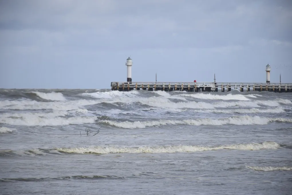 Ruwe zee met golven en een pier met twee vuurtorens onder een bewolkte lucht.